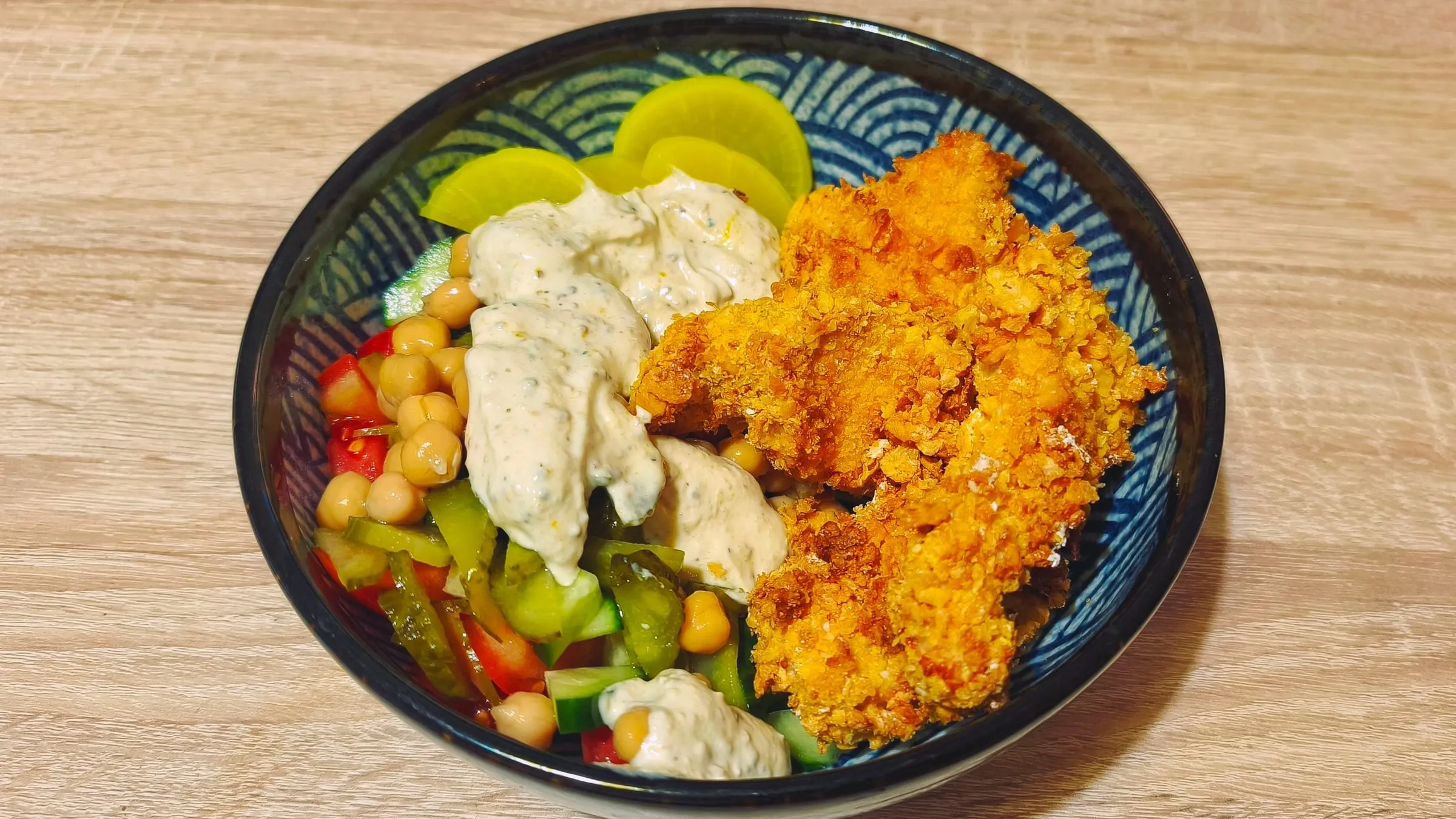 Un bol avec des aiguillettes de poulet panées et dorées, une salade de tomates, concombre, cornichons et pois chiche, une sauce crémeuse et des tranches de cornichon en décoration.