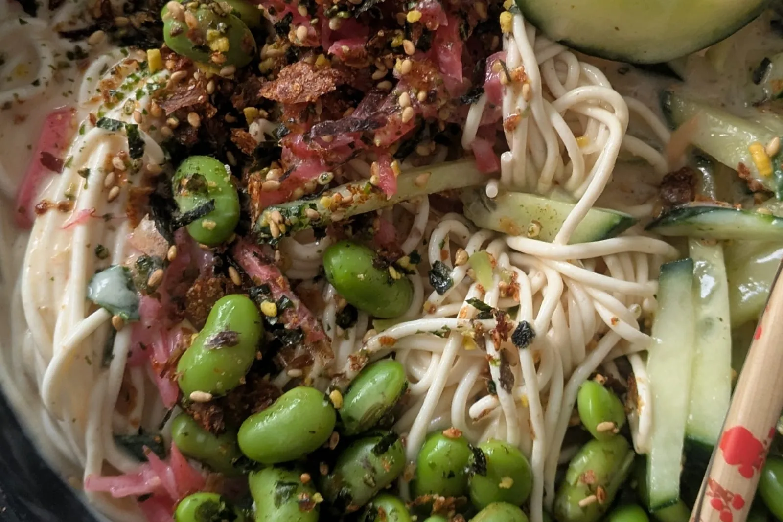 Un bol de nouilles froides nappées d'une soupe crémeuse claire, garni d'edamames, de concombre en lamelles, de gingembre rose et de furikake, avec des baguettes en bois posées sur le bord.