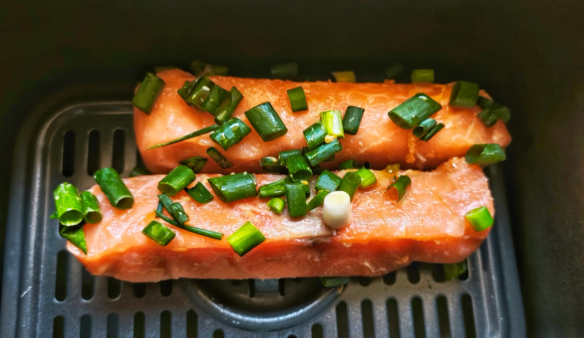 Deux pavés de saumon marinés couverts d'oignons nouveaux émincés, posés dans le panier de l'airfryer avant cuisson.