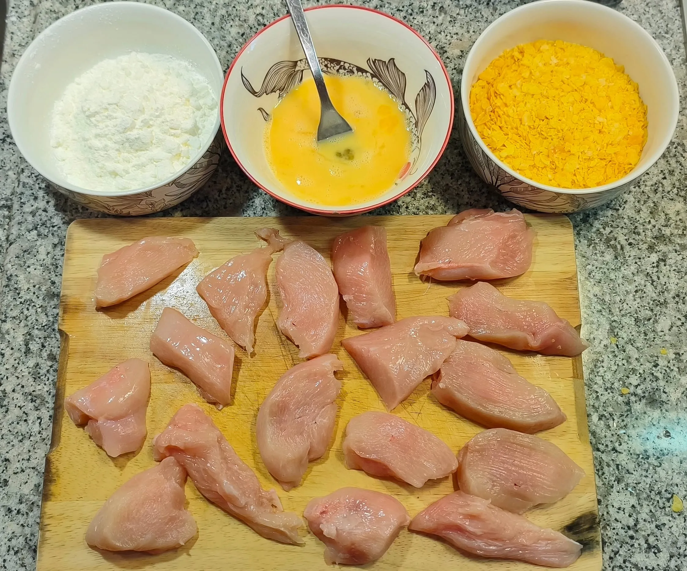 Trois bols vus de haut, un de maïzena, un d'œufs battus, un de corn flakes émiettés, posés au dessus d'une planche en bois avec des morceaux de poulet crus coupés en pièces égales.