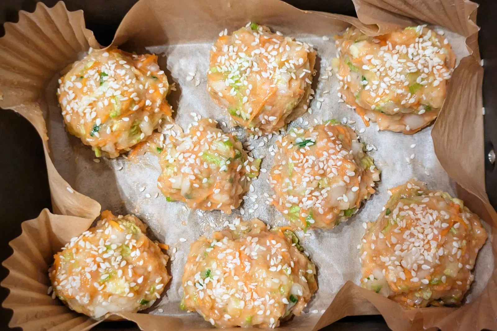 Huit boulettes de poulet et légumes enrobées de graines de sésame, disposées sur du papier sulfurisé dans un bac de airfryer.