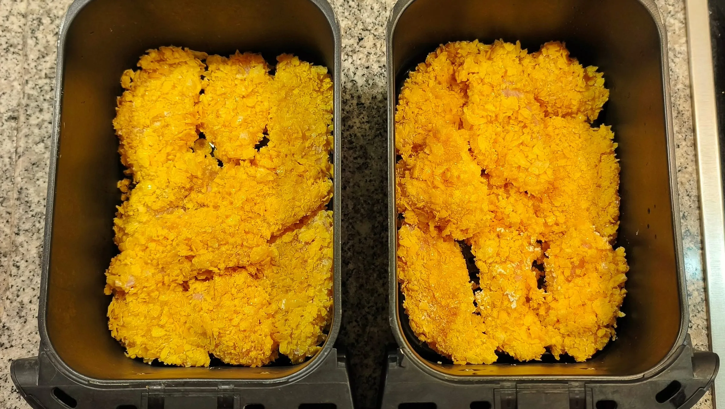 Vue de haut sur les deux bacs d'un airfryer double, chacun rempli de pièces de poulet panées aux corn flakes, dorées après cuisson.